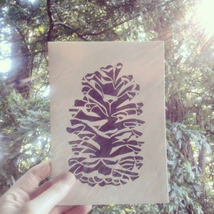 May include: A black and white print of a pine cone on a light brown card.
