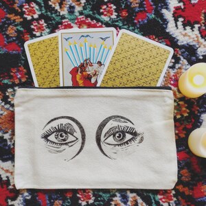 Op de afbeelding: Een witte canvas tas met een ritssluiting en twee zwarte ogen bedrukt op de voorkant. De tas is gevuld met tarotkaarten, waaronder een kaart met een gele achtergrond en een ontwerp van zwaarden en een andere kaart met een blauwe achtergrond en een ontwerp van mensen die zwaarden vasthouden.