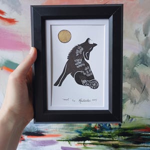 May include: Black framed art print featuring a howling wolf silhouette with a gold moon on a white background. The print is titled "Howl" and is numbered 1/10. The artwork is held by a hand.