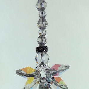 Peut inclure: Un attrape-soleil en cristal transparent avec un motif de fleur. Il est composé de perles de cristal facettées et d'un pendentif en forme de fleur. La fleur a six pétales aux bords irisés. L'attrape-soleil est suspendu à un fil noir.