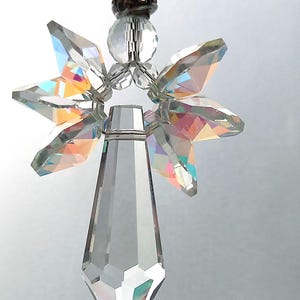 May include: A clear crystal angel ornament with iridescent wings and a faceted body. The angel has a star-shaped halo and is suspended by a string of clear beads. The ornament is designed to catch and reflect light.