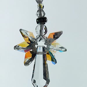 Könnte beinhalten: Ein klarer Kristallengel-Ornament mit facettierten Flügeln und einem tropfenförmigen Körper. Die Flügel zeigen einen Regenbogen von Farben. Das Ornament wird von einer Schnur aus klaren und grauen Perlen gehalten.