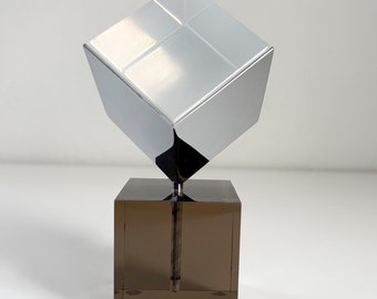 Könnte beinhalten: Eine Chrom-Würfelskulptur auf einem braunen quadratischen Sockel. Der Würfel ist reflektierend und scheint aus Metall zu sein.
