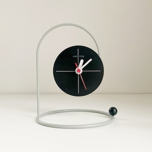 Puede incluir: Un reloj blanco y negro con un segundero rojo, montado en un soporte de metal gris. La esfera del reloj dice "AIKENTIME".