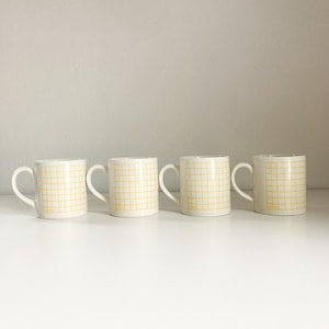 May include: Four white ceramic mugs with yellow grid patterns. The mugs have a handle on the right side.
