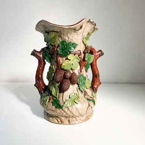May include: A ceramic vase with a tree trunk design. The vase is decorated with brown, green, and tan colours, with sculpted grape clusters and leaves. It has two brown handles and a wide, wavy rim.