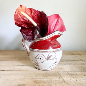 May include: A ceramic pitcher with a white base and a red glazed rim, featuring a hand-painted brown line drawing of a bird on the front. The pitcher contains two large anthurium flowers, one with deep red petals and one with light pink petals.