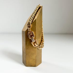 Puede incluir: Una pulsera de cadena dorada con un cierre decorativo se muestra sobre un expositor geométrico dorado. La pulsera presenta una cadena gruesa y texturizada y un cierre rectangular con un diseño único.