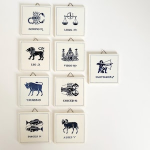 May include: A collection of square, white tiles featuring navy blue zodiac symbols and names. Each tile has a metal hanger. The zodiac signs include Scorpio, Libra, Leo, Virgo, Taurus, Cancer, Pisces, Aries, and Sagittarius.