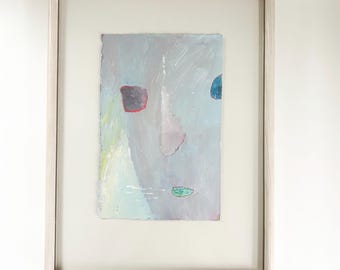 May include: Abstract painting in a white frame. The artwork features a face-like composition with gray, blue, red, and green shapes on a light gray background. The painting is framed with a white mat.