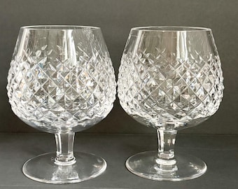 Puede incluir: Dos copas de brandy de cristal transparente con patrones de corte de diamante. Las copas tienen forma de cuenco redondeado, un tallo corto y una base circular. El diseño de cristal tallado refleja la luz. El fondo es gris oscuro.