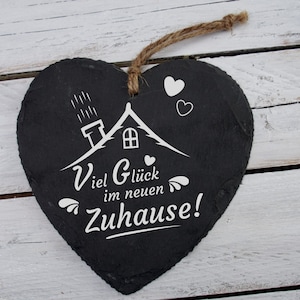 May include: Black heart-shaped slate sign with white lettering that reads "Viel Gl&uuml;ck im neuen Zuhause!"  The sign features a small white house with a chimney and a few raindrops above it. There are also a few small white hearts around the house.