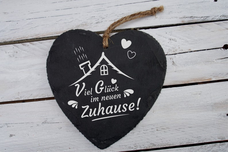 May include: Black heart-shaped slate sign with white lettering that reads "Viel Gl&uuml;ck im neuen Zuhause!"  The sign features a small white house with a chimney and a few raindrops above it. There are also a few small white hearts around the house.