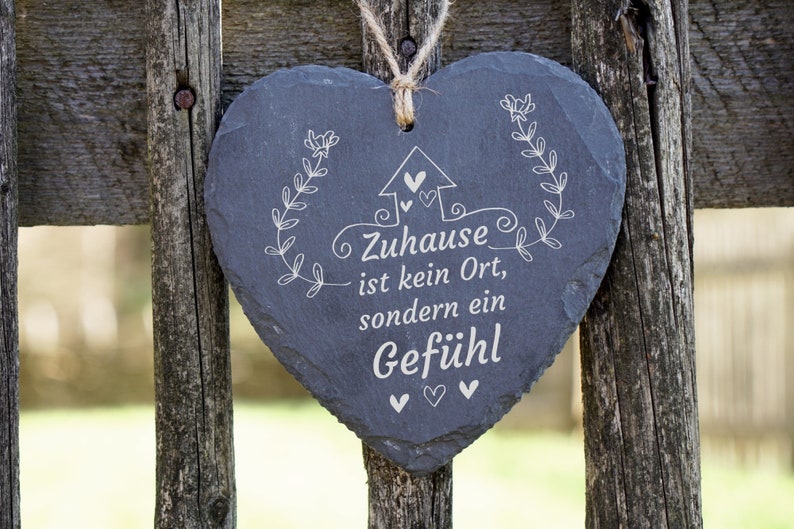 May include: A gray heart-shaped slate sign with white lettering that reads "Zuhause ist kein Ort, sondern ein Gef&uuml;hl" (Home is not a place, but a feeling). The sign is hanging on a wooden fence.