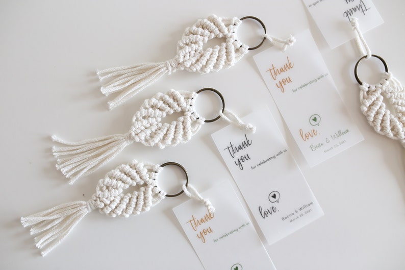 May include: Three white macrame keychains with tassels and a metal ring. Each keychain is attached to a white card with the text "Thank you for celebrating with us" and "love" with a heart icon. The cards also include the names "Bacis & Wilham" and the date "March 25, 2023".