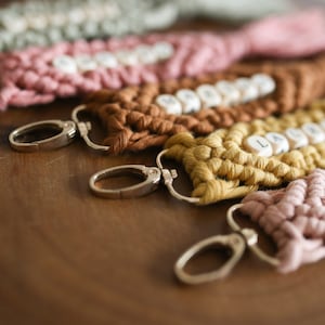Puede incluir: Cinco llaveros de macram&eacute; de colores con anillas de plata. Cada llavero tiene un nombre diferente escrito con cuentas de letras de madera. Los nombres son Emil, Lauryn, Maggie, Lily y un cuarto nombre que no es visible.