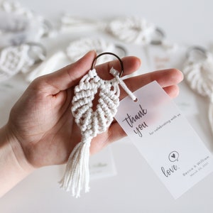 May include: White macrame keychain with a tassel, attached to a small, clear, plastic tag with the text "thank you for celebrating with us love. Becca & William March 20, 2021"