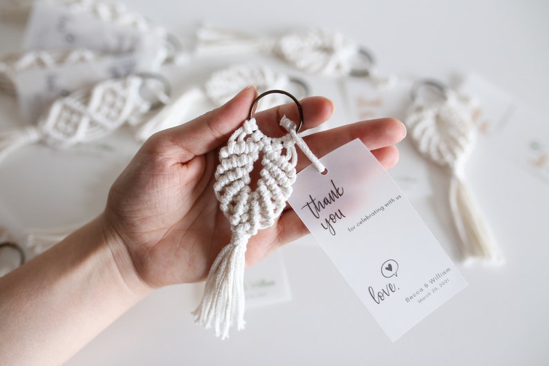 May include: White macrame keychain with a tassel, attached to a small, clear, plastic tag with the text "thank you for celebrating with us love. Becca & William March 20, 2021"
