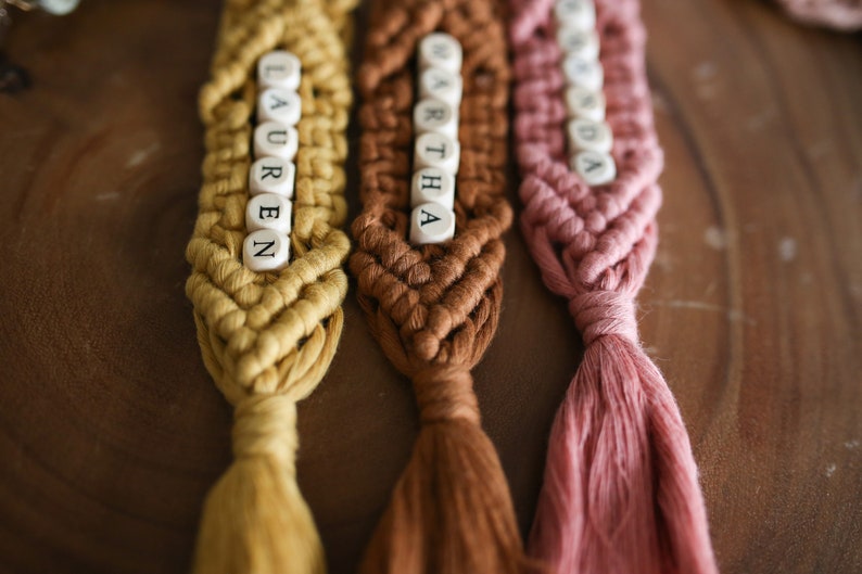 Puede incluir: Tres llaveros de macram&eacute; con borlas en amarillo, marr&oacute;n y rosa. Cada llavero tiene cuentas de letras blancas que deletrean los nombres "Lauren", "Martha" y "Lynda".