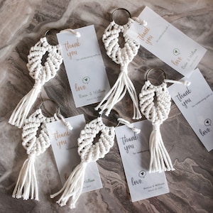 May include: A set of six white macrame keychains with gold key rings and a small white tag attached to each keychain. The tags have the text "Thank you" and "Love" printed on them.