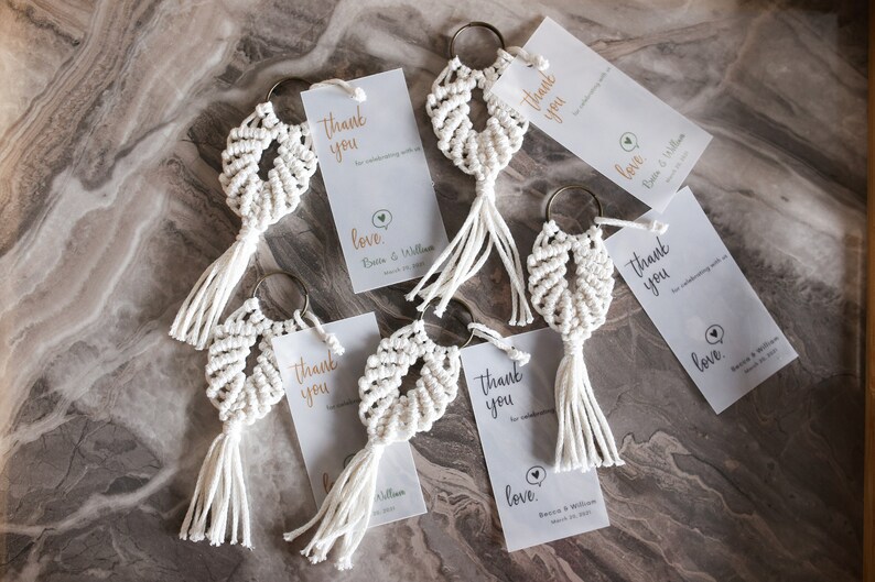 May include: A set of six white macrame keychains with gold key rings and a small white tag attached to each keychain. The tags have the text "Thank you" and "Love" printed on them.