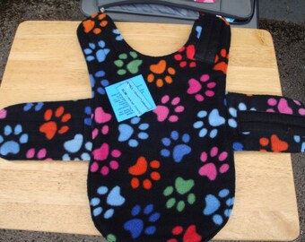 Könnte beinhalten: Ein schwarzer Fleece-Hundemantel mit einem Pfotenabdruckmuster in verschiedenen Farben, darunter Pink, Blau, Grün und Orange. Der Mantel hat einen Klettverschluss und einen schwarzen Riemen über dem Rücken. Das Etikett lautet "SMALL - EXTRA SMALL - 10-12 inch chest".