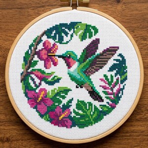 May include: A cross-stitch embroidery featuring a hummingbird, pink hibiscus flowers, and green leaves, set within a round wooden hoop. The detailed design uses various colours and is stitched on white fabric. The hoop is secured with a brass screw.