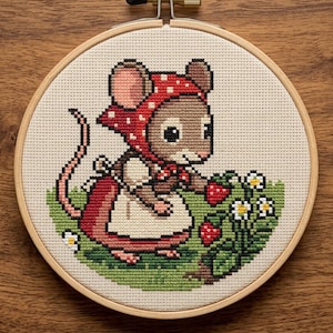 May include: Cross-stitch embroidery of a mouse wearing a red polka-dot headscarf and apron, picking strawberries. The mouse is surrounded by green grass, white flowers, and red strawberries. The embroidery is framed in a wooden hoop.