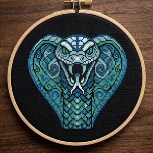 May include: A cross-stitch embroidery of a cobra in a wooden hoop. The cobra is depicted in shades of blue and teal against a black background. The embroidery hoop is made of wood and has a brass screw at the top.