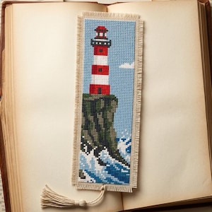 Könnte beinhalten: Ein rechteckiges Kreuzstich-Lesezeichen mit einem rot-weißen Leuchtturm auf einer dunkelgrünen Klippe über blau-weißen Wellen. Der Stoff hat einen ausgefransten Rand und eine hellbeige Quaste, die unten in der Mitte befestigt ist.