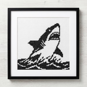 Peut inclure: Image d'une broderie au point de croix en noir et blanc représentant un requin sortant de l'eau, encadrée dans un cadre noir. Le requin a la gueule ouverte, montrant ses dents. L'œuvre est sur fond blanc.