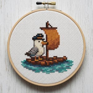 Könnte beinhalten: Kreuzstich-Stickerei in einem Holzreifen. Das Design zeigt eine Möwe mit Matrosenmütze auf einem kleinen Boot mit orangefarbenem Segel. Das Wasser ist in Blau- und Grüntönen gehalten. Der Hintergrund ist weiß.