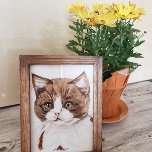 May include: A framed watercolor painting of a brown and white kitten with yellow eyes. The kitten is looking directly at the viewer. The frame is brown wood.
