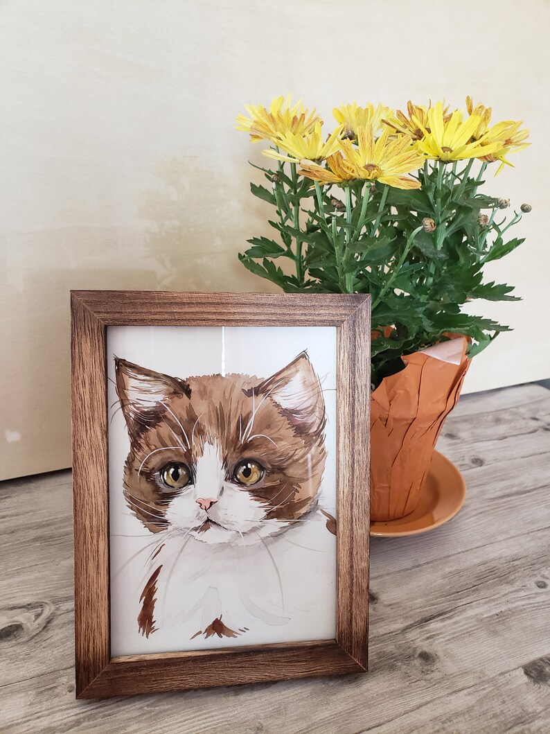 May include: A framed watercolor painting of a brown and white kitten with yellow eyes. The kitten is looking directly at the viewer. The frame is brown wood.