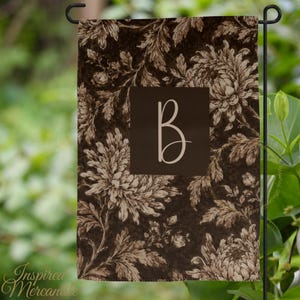 May include: A rectangular garden flag featuring a dark brown background with a repeating cream-colored floral pattern of chrysanthemums and leaves. A solid dark brown square is centered on the flag, displaying a large, cream-colored letter B.