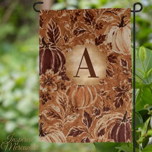 May include: A rectangular garden flag featuring a repeating pattern of pumpkins and autumn leaves in shades of brown, tan, and cream. A central light-colored scalloped plaque displays the capital letter A in a dark brown serif font.