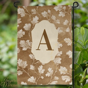 May include: A rectangular garden flag featuring a brown background with a repeating white oak leaf pattern. In the center, a cream-colored scalloped plaque displays a large, bold, dark brown capital letter A in a serif typeface.