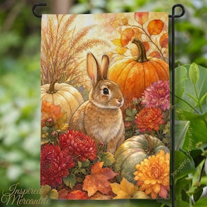 May include: A rectangular garden flag featuring a brown rabbit centered among orange and green pumpkins, vibrant red and yellow chrysanthemum flowers, and golden wheat stalks. The scene is set against a backdrop of autumn leaves in shades of orange.
