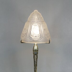 May include: A vintage Art Deco table lamp with a frosted glass shade and a brass base. The shade is shaped like a bell and features floral and geometric designs. The brass base has an octagonal shape with intricate detailing. The lamp is lit, casting a warm glow.