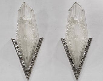 May include: A pair of silver and frosted glass wall sconces with a geometric design. The sconces have a pointed, triangular shape and feature a decorative, ornate border.