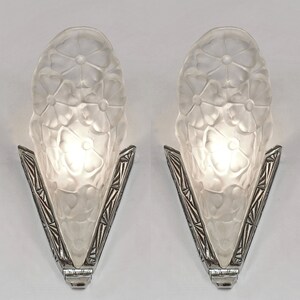 May include: Pair of silver wall sconces with frosted glass shades featuring a floral design. The shades are shaped like an elongated teardrop and have a textured surface.
