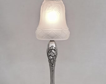 May include: A table lamp featuring a bell-shaped, frosted glass shade with embossed floral patterns and a diamond-grid base. The lamp has a metallic silver-toned stand with intricate floral relief carvings and a wide, circular, decorative base.