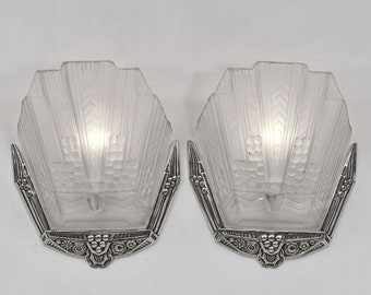 May include: Pair of silver and frosted glass Art Deco wall sconces with geometric designs. The sconces have a fan-shaped design and are accented with ornate details.