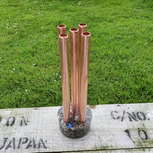 May include: A copper pipe flower vase with five openings, filled with small stones and shells. The vase is sitting on a wooden surface with the words "CANO" and "JAPAN" written on it.