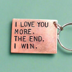 Può includere: Un portachiavi rettangolare color rame con le parole "I LOVE YOU MORE. THE END. I WIN." in lettere nere. Il portachiavi è attaccato a un anello diviso color argento. Lo sfondo è di colore verde acqua.