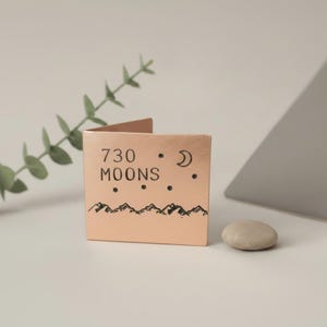 May include: A small, copper-coloured metal card holder with the words "730 MOONS" and a mountain range illustration. The design includes a crescent moon and stars. A small, smooth stone rests beside it.