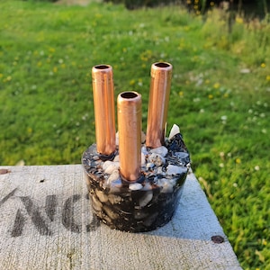 Könnte beinhalten: Eine schwarz-weiß gesprenkelte Keramikvase mit drei herausstehenden Kupferrohren. Die Vase steht auf einer Holzoberfläche.