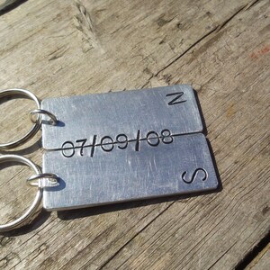 May include: Close-up of a pair of silver metal keychains. Each keychain has a rectangular tag engraved with a date, '07/09/08', and a compass direction, 'N' and 'S'. The keychains are linked together and rest on a rustic wooden surface.Personalised Anniversary Keyring Set Initial & Date Keychain Christmas Birthday Engagement Pair Keychain Couples