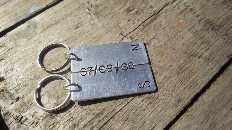May include: Close-up of a pair of silver metal keychains. Each keychain has a rectangular tag engraved with a date, '07/09/08', and a compass direction, 'N' and 'S'. The keychains are linked together and rest on a rustic wooden surface.Personalised Anniversary Keyring Set Initial & Date Keychain Christmas Birthday Engagement Pair Keychain Couples