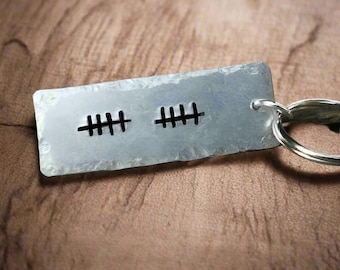 May include: A rectangular, silver-colored metal keychain with two sets of tally marks etched into the surface. The keychain is attached to a silver key ring. The background is a blurred, brown wooden surface.